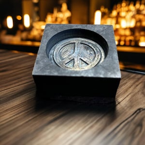 May include: A black square-shaped ashtray with a peace sign design in the centre. The ashtray is made of a textured material and has a dark grey finish.