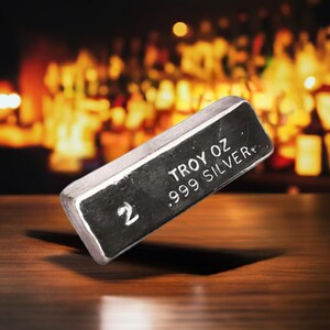 May include: A 2 troy ounce silver bar with the inscription "TROY OZ .999 SILVER" on a wooden surface.