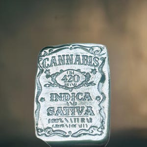May include: A silver metal charm with the words "Cannabis 420 Indica Sativa 100% Natural Grown in Cali" engraved on it.