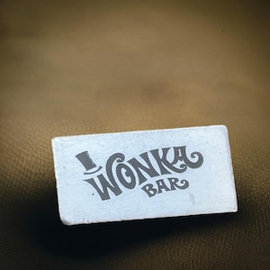 May include: A rectangular, light-colored Wonka Bar sign with the words "Wonka Bar" in a stylized font. The top hat logo is above the word "Wonka". The sign is set against a brown background.