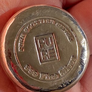 Puede incluir: Una barra de lingotes de plata con las palabras "Pure Scottish Silver 999 Fine Silver" y un logotipo que dice "Pure" en un cuadrado.