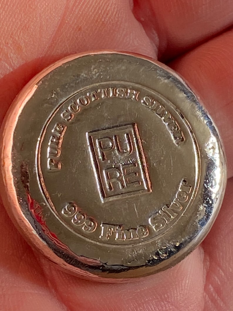 Puede incluir: Una barra de lingotes de plata con las palabras "Pure Scottish Silver 999 Fine Silver" y un logotipo que dice "Pure" en un cuadrado.