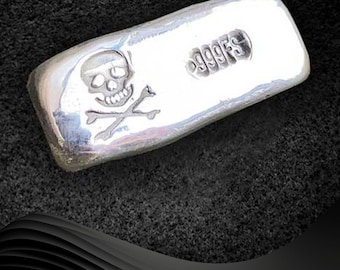 Könnte beinhalten: Ein Silberbarren mit einem Totenkopf- und Knochenkreuz-Design und der Aufschrift "999.95" auf der Seite.