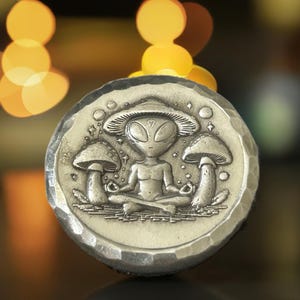 May include: A silver-toned metal coin with a detailed design of an alien meditating with mushrooms around it. The alien is wearing a mushroom cap and has large eyes. The coin has a textured surface.