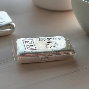 May include: A silver bar with the words "100 Grams" and "Pure" engraved on it. The bar also has a cross-shaped design.