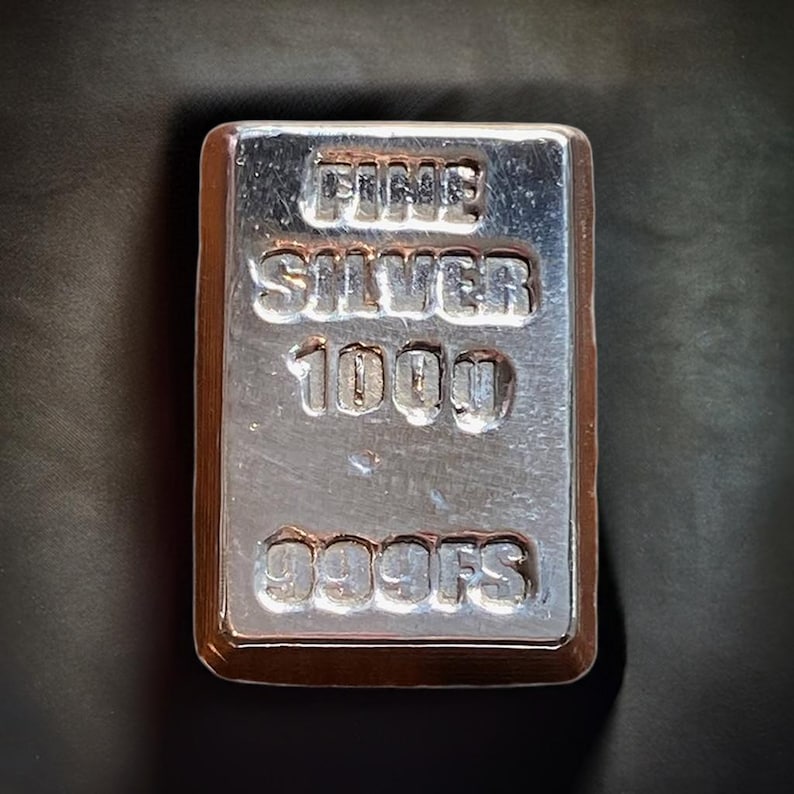 Pode incluir: Uma barra de prata com as palavras "FINE SILVER 1000 999FS" gravadas em relevo.