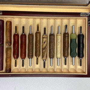 May include: A wooden box with a velvet lining containing 12 different wood burning tools. The tools are arranged in a row and have a variety of wood grain patterns.
