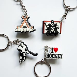 May include: Four hockey-themed keychains with metal rings and chains. One features crossed hockey sticks and a puck, one a goalie in a net, one a hockey player in motion, and one a white rectangular charm with the text I LOVE HOCKEY.