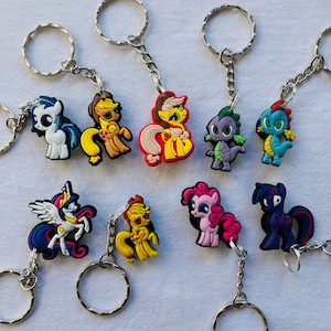 Könnte beinhalten: Ein Set mit zehn bunten Schlüsselanhängern mit Cartoon-Charakteren, die My Little Pony und Drachenfiguren darstellen. Die Schlüsselanhänger sind aus Gummi und haben silberne Schlüsselringe.