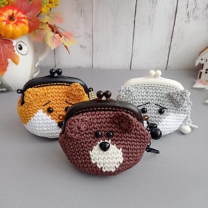 May include: Three small, round, crocheted coin purses shaped like animal faces. One is orange and white, one is brown and white, and one is gray and white. Each purse features a dark plastic kiss-lock clasp, small ears, and black bead eyes.