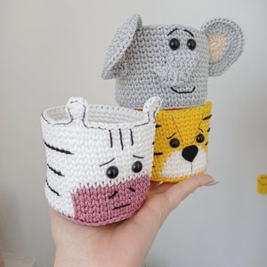 May include: Three handmade crochet animal-themed containers. The top container is gray and shaped like an elephant. The middle container is yellow and white, resembling a tiger. The bottom container is white with black stripes, resembling a zebra.