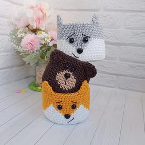 May include: Three stacked crochet baskets shaped like animal faces. The top basket is gray and white with pointed ears, the middle is dark brown with a tan muzzle, and the bottom is orange and white with pointed ears. Each has black bead eyes and noses.