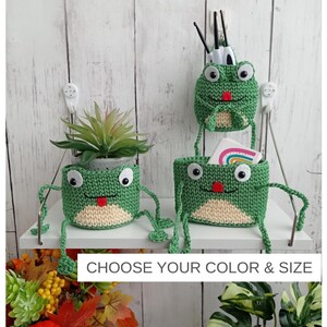 May include: Three green crochet frog-shaped containers with cream-colored bellies, large white eyes, and red tongues. One hangs on a wall, while two sit on a shelf. The text CHOOSE YOUR COLOR & SIZE is printed in black on a white rectangular background.