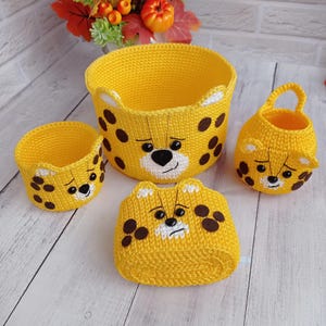 May include: A set of four handmade yellow crochet baskets with a cheetah face design. The baskets vary in size, with brown spots and white accents. The largest basket has handles. The cheetah face includes a black nose and sad eyes.