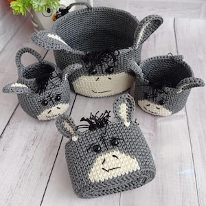 Puede incluir: Un conjunto de cuatro cestas de ganchillo con temática de burro, grises y blancas. Las cestas varían en tamaño, la más grande tiene un asa. Cada cesta presenta una cara de burro con ojos negros, hocico blanco y lana negra para la crin.