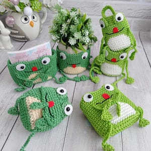 May include: A collection of green crocheted frog-themed organizers and containers. Each item features large white eyes with black pupils, a small red tongue, and a cream-colored belly. The set includes a rectangular basket, a plant pot cover, and pouches.