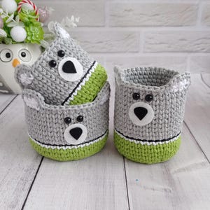 May include: Two cylindrical crochet baskets featuring a gray upper section and a lime green base. Each basket has a bear-like face with black plastic eyes, a white felt muzzle, and a black felt nose. The baskets have small ears on the top rim.