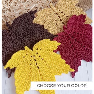 Pode incluir: Quatro bases para copos em croché com formato de folha nas cores castanho escuro, amarelo mostarda, bege claro e bordeaux. Cada folha apresenta um caule central e nervuras cosidas. O texto CHOOSE YOUR COLOR está num retângulo branco.