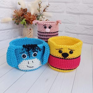 May include: Three crocheted baskets with cartoon animal faces. The baskets are blue, yellow, and pink. The blue basket has a donkey face, the yellow basket has a bear face, and the pink basket has a pig face.