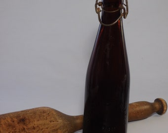 May include: A dark brown glass bottle with a wire closure and a white plastic stopper. The bottle is sitting on a wooden surface next to a wooden mallet.