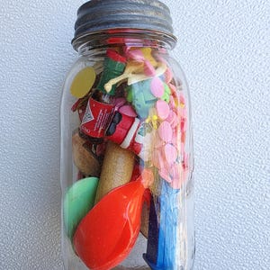 May include: A clear glass jar filled with a variety of small, colorful objects. The jar has a silver lid and contains items like miniature toys, wooden pieces, and plastic shapes. The contents are a mix of red, green, blue, and yellow.