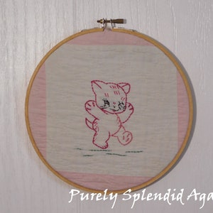 May include: A pink and white embroidered hoop art featuring a pink kitten with black eyes. The kitten is standing on its hind legs with its front paws raised.