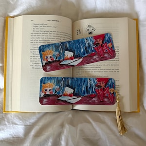 May include: Two illustrated bookmarks with a gold tassel rest inside an open book. The bookmarks feature a cartoon character with red and blue accents. The book has yellow covers and is open to page 24.