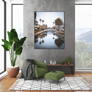 May include: A green armchair with a gray throw blanket sits in a living room with a large window overlooking a lush green landscape. A framed photograph of a canal with palm trees and a bridge hangs on the wall behind the chair. A small green ottoman sits in front of the chair.