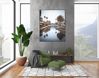 May include: A green armchair with a gray throw blanket sits in a living room with a large window overlooking a lush green landscape. A framed photograph of a canal with palm trees and a bridge hangs on the wall behind the chair. A small green ottoman sits in front of the chair.