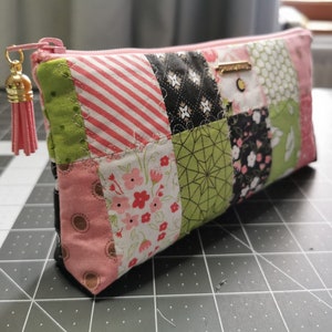 Peut inclure: Une pochette zippée en patchwork avec une fermeture éclair et un pompon roses. La pochette est composée de divers carrés de tissu aux motifs roses, verts, noirs et blancs, dont des rayures, des motifs floraux et géométriques. Une petite étiquette dorée est attachée.