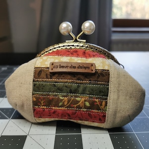 Peut inclure: Un porte-monnaie fait à la main avec un motif patchwork. Le sac à main est doté d'un fermoir en métal avec des accents de perles. Le patchwork comprend divers tissus dans les tons de rouge, vert et marron, avec un tissu beige semblable à du lin sur les côtés. Une petite étiquette en cuir indique "au bout des doigts".