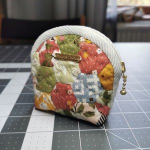 Peut inclure: Un petit porte-monnaie fait à la main avec un motif patchwork. Le sac présente une variété de tissus à motifs floraux et géométriques dans des tons de rouge, vert, jaune et bleu. Il a une bordure rayée et une fermeture éclair dorée.