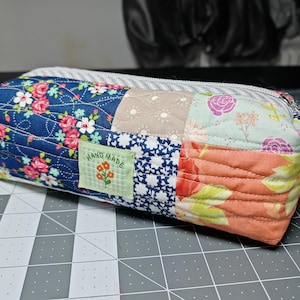 Może przedstawiać: Prostokątny, pikowany piórnik z patchworkowym wzorem. Etui posiada różnorodne tkaniny w kwiatowe wzory w odcieniach niebieskiego, różowego, brzoskwiniowego i beżowego. Mała zielona metka z napisem "HAND MADE". Etui posiada srebrny zamek błyskawiczny.