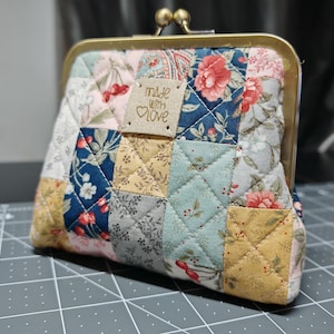 Può includere: Un portamonete patchwork con chiusura in ottone. La borsa è realizzata con vari quadrati di tessuto con motivi floreali e geometrici, tra cui rosa, blu e giallo. Una piccola etichetta recita "made with love."