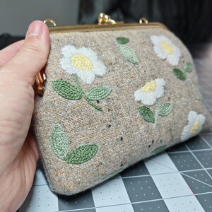 Puede incluir: Un bolso de mano de tela beige con cierre dorado, con flores blancas bordadas con centros amarillos y hojas verdes. El bolso se sostiene en una mano, mostrando el detallado diseño floral.