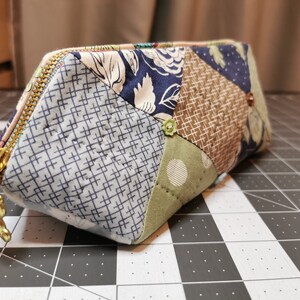 Könnte beinhalten: Ein Patchwork-Stoff-Reißverschlusstasche mit einem goldfarbenen Reißverschluss und einem Anhänger. Die Tasche besteht aus verschiedenen gemusterten Stoffen in Blau-, Grün- und Brauntönen. Der Anhänger ist ein kleiner Bär. Die Tasche ist etwa 15 cm lang.