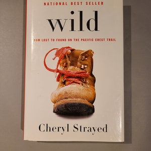 May include: A book cover with the title "wild" in bold, black font. The subtitle is "From Lost to Found on the Pacific Crest Trail". The cover features a close-up of a brown hiking boot with red laces. The author's name, "Cheryl Strayed", is printed in black at the bottom of the cover.