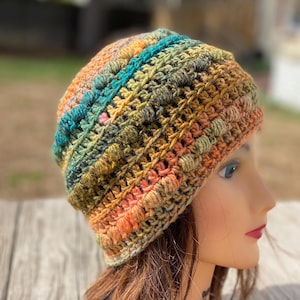 May include: A crochet beanie featuring a variegated yarn pattern with horizontal bands of orange, teal, olive green, and brown. The hat is constructed with a textured stitch pattern that includes raised bobbles and openwork rows throughout the design.