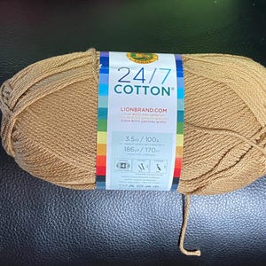 May include: A ball of Lion Brand 24/7 Cotton yarn in a warm, golden-brown shade. The label shows the brand name, yarn weight, and length, with a rainbow stripe. The yarn is 100g and 170m.