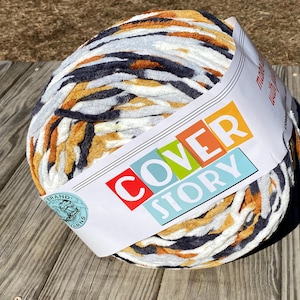 May include: A large ball of Cover Story brand yarn in a mix of colors including white, orange, gray, and black. The yarn is wrapped with a label that reads "COVER STORY" in colorful block letters. The yarn is sitting on a weathered wooden surface.