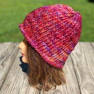 May include: A chunky knit beanie hat featuring a variegated yarn pattern in vibrant shades of magenta, deep purple, bright orange, and red. The hat is constructed with thick, textured stitches and has a rounded crown with a slightly folded brim edge.