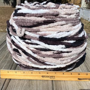 May include: A large ball of chunky yarn in shades of black, white, and various shades of brown. The yarn is thick and soft, perfect for knitting or crocheting projects. A wooden ruler is placed next to the yarn ball, measuring up to 11 inches.