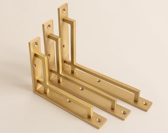 May include: Three L-shaped shelf brackets made of brushed gold-toned metal. Each bracket features a vertical mounting plate with pre-drilled screw holes and a horizontal support arm connected by a perpendicular brace, creating a geometric frame.