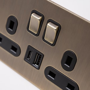 May include: A brushed antique brass electrical outlet with two toggle switches, two USB ports, and two standard outlets. The outlets and switches are black.