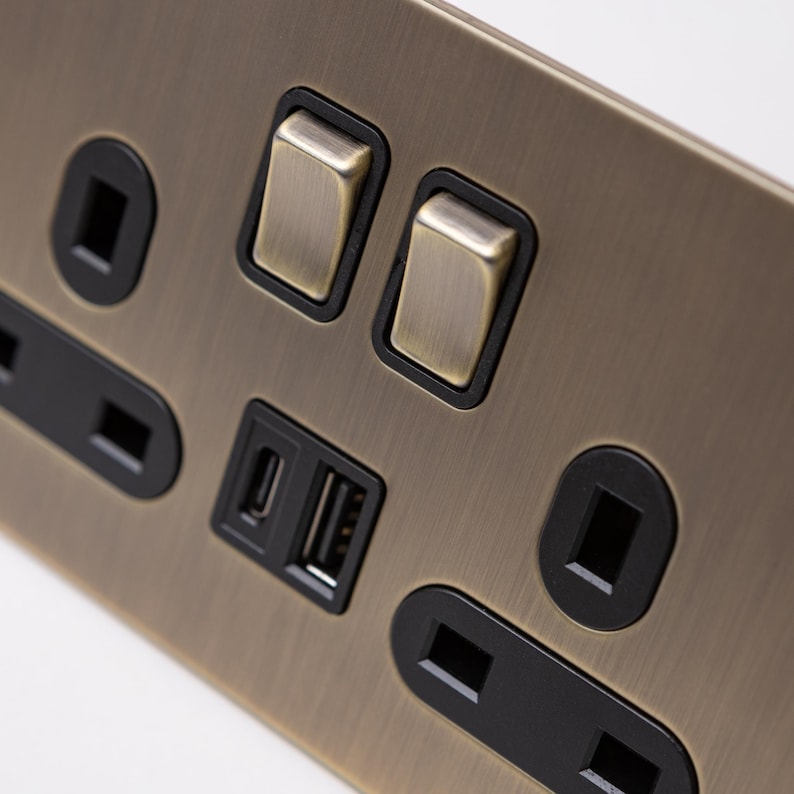May include: A brushed antique brass electrical outlet with two toggle switches, two USB ports, and two standard outlets. The outlets and switches are black.