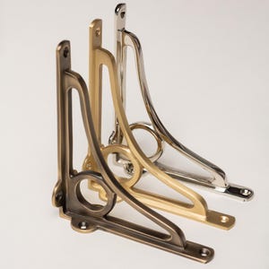 May include: Three metal shelf brackets in bronze, gold, and silver finishes. Each bracket has a decorative, curved design with a circular cutout and mounting holes for installation. The brackets are displayed against a white background.