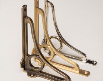 May include: Three L-shaped metal shelf brackets arranged in a row. Each bracket features a decorative circular cutout at the corner and mounting holes at the ends. The finishes include a dark bronze, a brushed gold, and a polished silver tone.