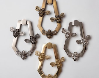 May include: Four metal door knockers arranged in a cluster, each featuring a hexagonal frame adorned with three bee figures. The knockers are finished in various metallic tones, including brushed brass, polished silver, and antique bronze finishes.
