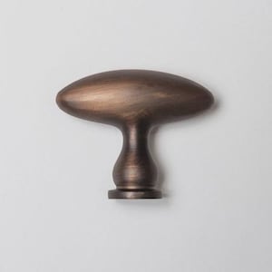 Peut inclure: Un bouton de meuble en forme de T avec une finition en bronze foncé lisse. Le bouton présente une partie supérieure ovale horizontale allongée qui se rétrécit vers une base arrondie en forme de piédestal. La surface métallique est nuancée.