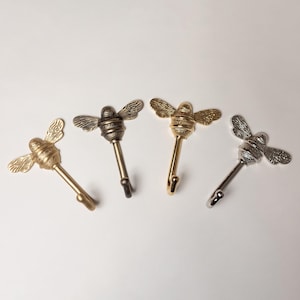May include: Four metal wall hooks, each featuring a bee-shaped top with textured wings and a striped body. The hooks are arranged in a row, displaying four distinct metallic finishes: matte gold, antique brass, polished gold, and polished silver.